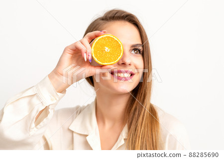 Portrait of a cheerful caucasian young woman covering eye with an orange slice wearing a white shirt over a white background 88285690
