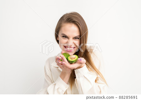 Portrait of a lovely smiling young brunette caucasian woman wearing the white shirt with long hair holding and showing avocado, standing isolated over white background Portrait of a lovely smiling young brunette caucasian woman wearing the white shirt with long hair holding and showing avocado, standing isolated over white background 88285691