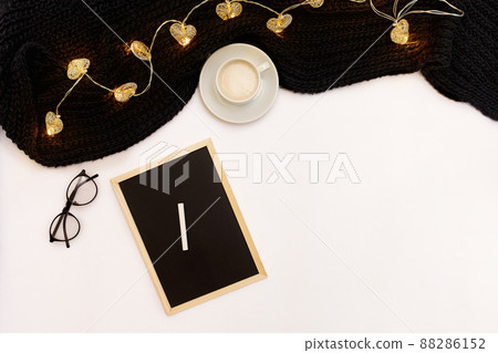 A cup of coffee, a small black wooden board and a white chalk with new years goals. Scarf and Christmas lights on white background. 88286152