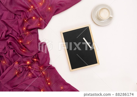 A cup of coffee, a small black wooden board and a white chalk with new years goals. Scarf and Christmas lights on white background. A cup of coffee, a small black wooden board and a white chalk with new years goals. Scarf and Christmas lights on white background. 88286174