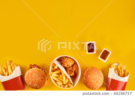 Fast food dish top view. Meat burger, potato chips and wedges. Take away composition. French fries, hamburger, mayonnaise and ketchup sauces on yellow background. Fast food dish top view. Meat burger, potato chips and wedges. Take away composition. French fries, hamburger, mayonnaise and ketchup sauces on yellow background. 88286757