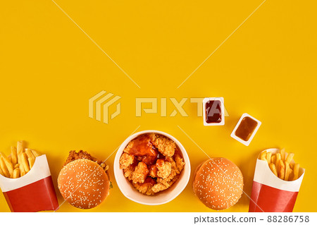 Fast food dish top view. Meat burger, potato chips and wedges. Take away composition. French fries, hamburger, mayonnaise and ketchup sauces on yellow background. Fast food dish top view. Meat burger, potato chips and wedges. Take away composition. French fries, hamburger, mayonnaise and ketchup sauces on yellow background. 88286758
