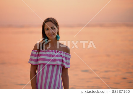 Tanned beautiful woman in dress with white and pink stripes. Close up and copy space. Orange sea or ocean at the sunset. Vacations concept Tanned beautiful woman in dress with white and pink stripes. Close up and copy space. Orange sea or ocean at the sunset. Vacations concept 88287160