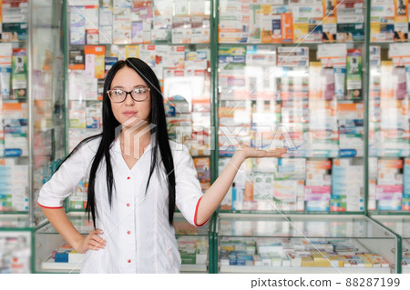 Pharmacy and pharmacist-seller. Female pharmacist with glasses posing on the background of shop Windows and pointing. Copy space Pharmacy and pharmacist-seller. Female pharmacist with glasses posing on the background of shop Windows and pointing. Copy space 88287199