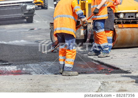 A group of road workers in orange overalls are repairing a section of the carriageway by rolling up fresh asphalt. 88287356
