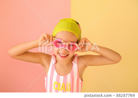 A child in a swimsuit and goggles and a swimming cap smiles sweetly looking at the camera. Girl swimmer Pink and yellow studio background. 88287769