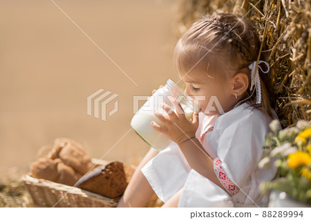 a healthy childhood in harmony with nature. happy little baby in folk clothes sits on a wheat field with a jar of fresh milk. the sun's rays are reflected in a jar of fresh, tasty milk 88289994