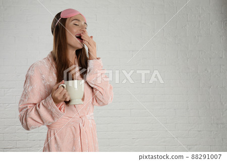 Beautiful young woman in bathrobe and eye sleeping mask yawning near white brick wall. Space for text 88291007