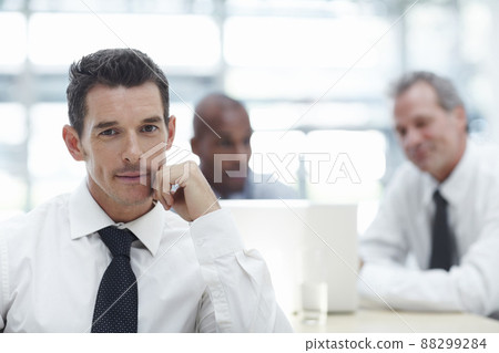 Thinking business. A handsome businessman sitting with his chin rested on his hand looking at the camera with two colleagues in the background working on a laptop. 88299284