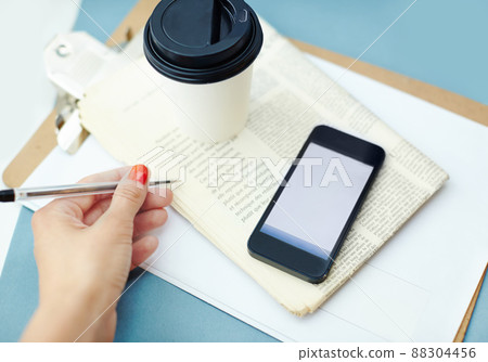 Take down this information. Disposable cup sitting ontop of a newspaper and clipboard, with a hand holding a pen and a smartphone. 88304456