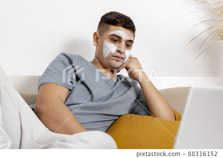 Young man with casual clothes and beauty mask on the face lying on bed with laptop computer and resting. Cosmetic for men, skin care. Man reading news in internet or watching movie. Relaxing at home. 88316152
