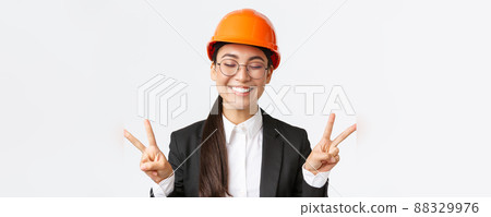 Close-up of cheerful successful female asian engineer, construction architect in safety helmet and business suit showing peace signs and smiling kawaii, standing white background Close-up of cheerful successful female asian engineer, construction architect in safety helmet and business suit showing peace signs and smiling kawaii, standing white background 88329976