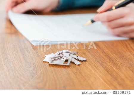 Close up of key with keychain lies on the table. Defocused hands signing a contract at the background. Concept of purchase of real estate and mortgage Close up of key with keychain lies on the table. Defocused hands signing a contract at the background. Concept of purchase of real estate and mortgage 88339171