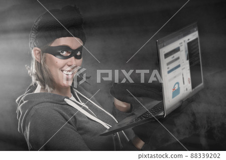 The concept of cybercrime and hacking. Portrait of a woman in a black hat, gloves and mask holding a laptop with a browser window about unlocking the password. Black background and light 88339202