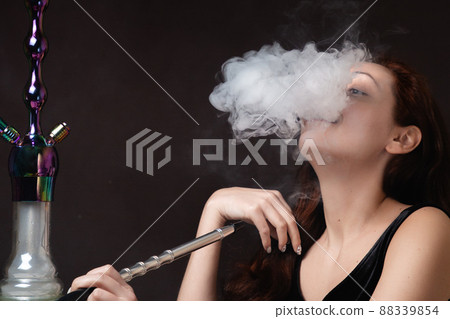 A beautiful young woman in a black dress smokes a hookah and lets out a cloud of steam. Face hidden in a cloud of steam. Close up A beautiful young woman in a black dress smokes a hookah and lets out a cloud of steam. Face hidden in a cloud of steam. Close up 88339854
