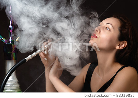 A beautiful young woman in a black dress smokes a hookah and lets out a cloud of steam. The atmosphere of relaxation and pleasure. Close up A beautiful young woman in a black dress smokes a hookah and lets out a cloud of steam. The atmosphere of relaxation and pleasure. Close up 88339855