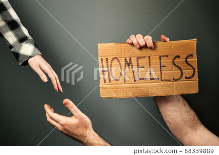 A woman extends a helping hand to a man holding a cardboard box with the text homeless. Dark background of a greenish hue. The concept of a social problem with people without a home and vagabonds 88339889