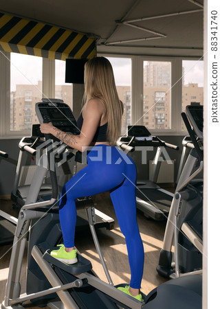 Side view of young sport woman in modern gym. Athletic girl trains on an elliptical trainer. Side view of young sport woman in modern gym. Athletic girl trains on an elliptical trainer. 88341740