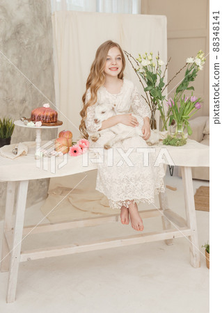 The girl is sitting at the Easter table with cakes, spring flowers and quail eggs and holding a white kid in her arms. Happy Easter celebration The girl is sitting at the Easter table with cakes, spring flowers and quail eggs and holding a white kid in her arms. Happy Easter celebration 88348141