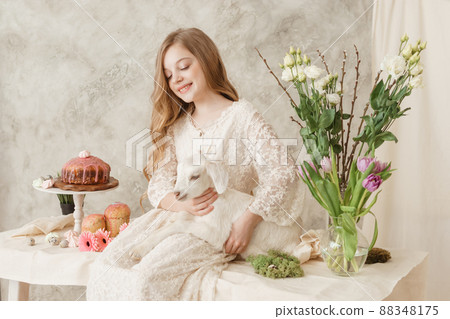 The girl is sitting at the Easter table with cakes, spring flowers and quail eggs and holding a white kid in her arms. Happy Easter celebration 88348175