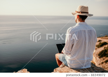 Digital nomad, man in the hat, a businessman with a laptop sits on the rocks by the sea during sunset, makes a business transaction online from a distance. Remote work on vacation. 88349072