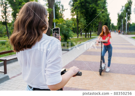 Two girl friends riding push e scooter and do selfie via their smartphones outdoors. Group gen z young people using electric scooter in city park Two girl friends riding push e scooter and do selfie via their smartphones outdoors. Group gen z young people using electric scooter in city park 88350256