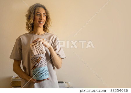 Happy young creative blonde female holding pile of hank reel ribbon yarn standing at home Happy young creative blonde female holding pile of hank reel ribbon yarn standing at home 88354086
