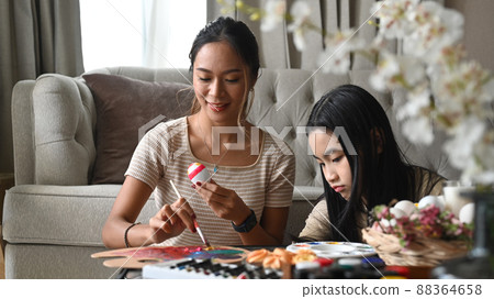 Happy mother and daughter enjoy painting eggs while preparing for Easter egg festival at home. 88364658