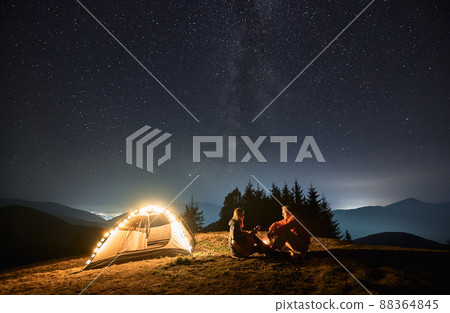 Woman with guitar in her hands sitting opposite man by campfire near their light shining tent. Romantic evening with loved one outdoors under starry sky in the mountains. Concept of night camping. 88364845