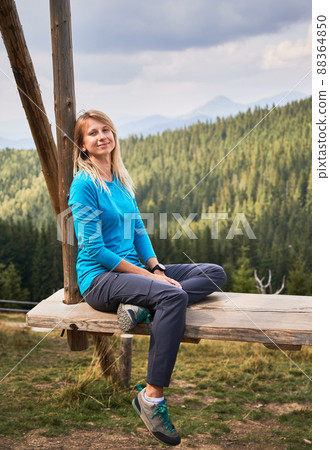 Portrait of pretty woman who sitting on edge of wooden seat, folding one leg on seat, leaning on side support with peaceful smile on face. Summer mountain landscape behind her. Portrait of pretty woman who sitting on edge of wooden seat, folding one leg on seat, leaning on side support with peaceful smile on face. Summer mountain landscape behind her. 88364850