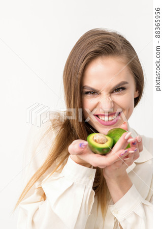 Portrait of a lovely smiling young brunette caucasian woman wearing the white shirt with long hair holding and showing avocado, standing isolated over white background Portrait of a lovely smiling young brunette caucasian woman wearing the white shirt with long hair holding and showing avocado, standing isolated over white background 88365956