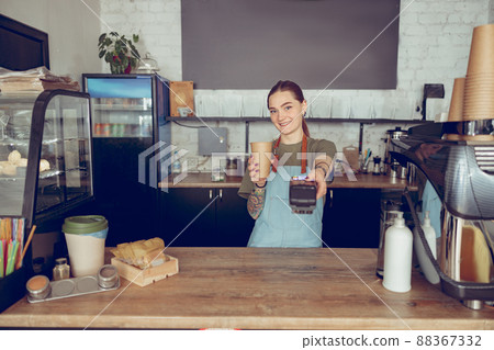Cheerful woman barista holding contactless card reader and coffee cup Cheerful woman barista holding contactless card reader and coffee cup 88367332