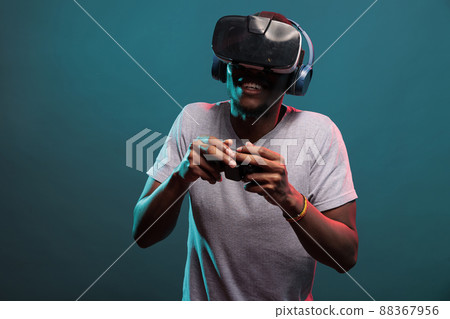 Modern gamer playing video game with joystick and vr headset, enjoying online play with futuristic 3d simulation in studio. Young man with virtual reality glasses holding controller. Modern gamer playing video game with joystick and vr headset, enjoying online play with futuristic 3d simulation in studio. Young man with virtual reality glasses holding controller. 88367956