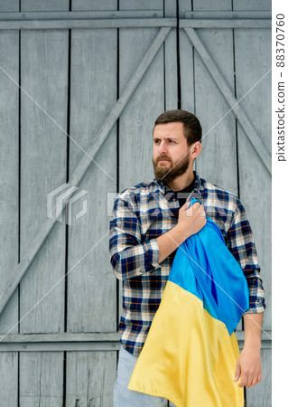Young man holding blue-yellow State Flag of Ukraine in right hand over heart on gray wooden fence background. Guy in a checkered shirt, vertical photo. Conceptual idea - with Ukraine in the heart. 88370760
