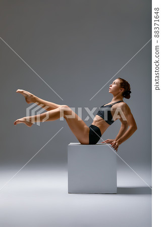 Flexible athletic woman in a sportswear doing splits in a fitness studio on a gray background. Stretching exercises with a cube Flexible athletic woman in a sportswear doing splits in a fitness studio on a gray background. Stretching exercises with a cube 88371648