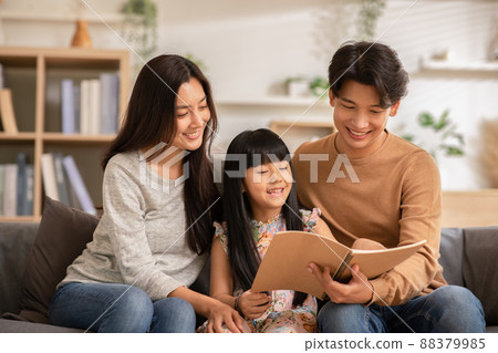 Happy Asian family sitting on the sofa together looking at daughter's note book. Parents are smiling, reading daughter book and proud of education progress. Watching book together, family relationship 88379985