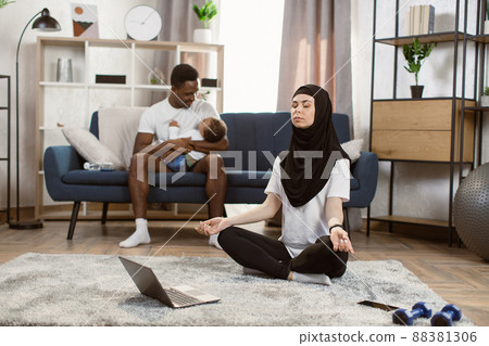 Arabian muslim woman in hijab meditating on mat in the living room using laptop Arabian muslim woman in hijab meditating on mat in the living room using laptop 88381306