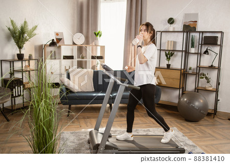 Fit woman running on the treadmill while drinking water from a bottle and doing her fitness exercise Fit woman running on the treadmill while drinking water from a bottle and doing her fitness exercise 88381410