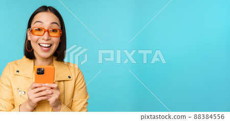Portrait of enthusiastic asian woman in sunglasses, using mobile phone, smiling and laughing, looking happy, holding smartphone, standing over blue background Portrait of enthusiastic asian woman in sunglasses, using mobile phone, smiling and laughing, looking happy, holding smartphone, standing over blue background 88384556