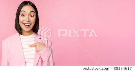 Portrait of young asian businesswoman with surprised, excited face expression, pointing finger at herself, standing in suit over pink background 88384617