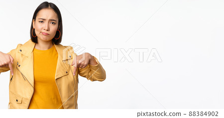 Portrait of disappointed asian woman pointing fingers down, grimacing and showing smth bad, standing over white background 88384902