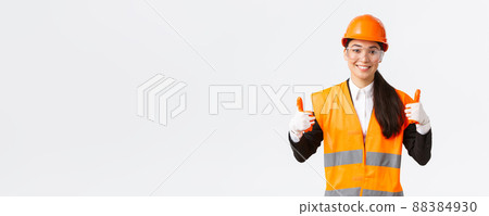 Satisfied smiling female asian chief construction engineer giving permission to enter building or entertprise after wearing safety clothing, glasses gloves and helmet, showing thumbs-up in approval 88384930