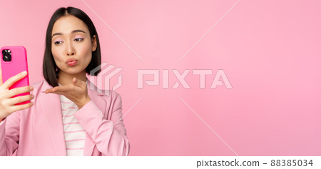Stylish asian businesswoman, girl in suit taking selfie on smartphone, video chat with mobile phone app, posing against pink studio background Stylish asian businesswoman, girl in suit taking selfie on smartphone, video chat with mobile phone app, posing against pink studio background 88385034