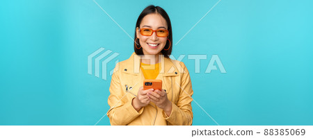 Smiling asian girl in sunglasses, using smartphone app, holding mobile phone, standing over blue background Smiling asian girl in sunglasses, using smartphone app, holding mobile phone, standing over blue background 88385069