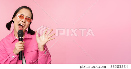 Happy beautiful asian girl singing with mic, using microphone, enjoying karaoke, posing against pink studio background Happy beautiful asian girl singing with mic, using microphone, enjoying karaoke, posing against pink studio background 88385284