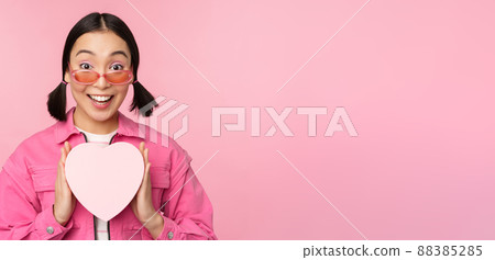 Beautiful asian girl smiling happy, showing heart gift box and looking excited at camera, standing over pink romantic background Beautiful asian girl smiling happy, showing heart gift box and looking excited at camera, standing over pink romantic background 88385285
