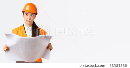 Thoughtful professional asian female construction manager, architect reading blueprints and looking away pondering, thinking, making decision while studying project plan, white background 88385286