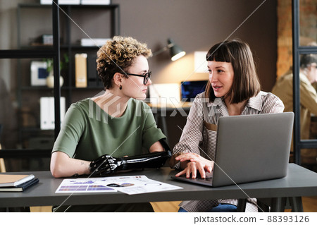Young businesswoman talking to employee with prosthetic arm at the table with laptop and documents at office 88393126