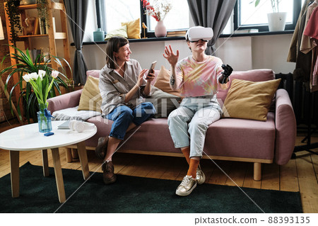 Girl with prosthetic arm in goggles playing virtual reality game with her friend while she using mobile phone on sofa in living room Girl with prosthetic arm in goggles playing virtual reality game with her friend while she using mobile phone on sofa in living room 88393135