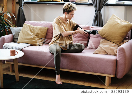 Young woman sitting on sofa in living room and putting on prosthetic arm Young woman sitting on sofa in living room and putting on prosthetic arm 88393189